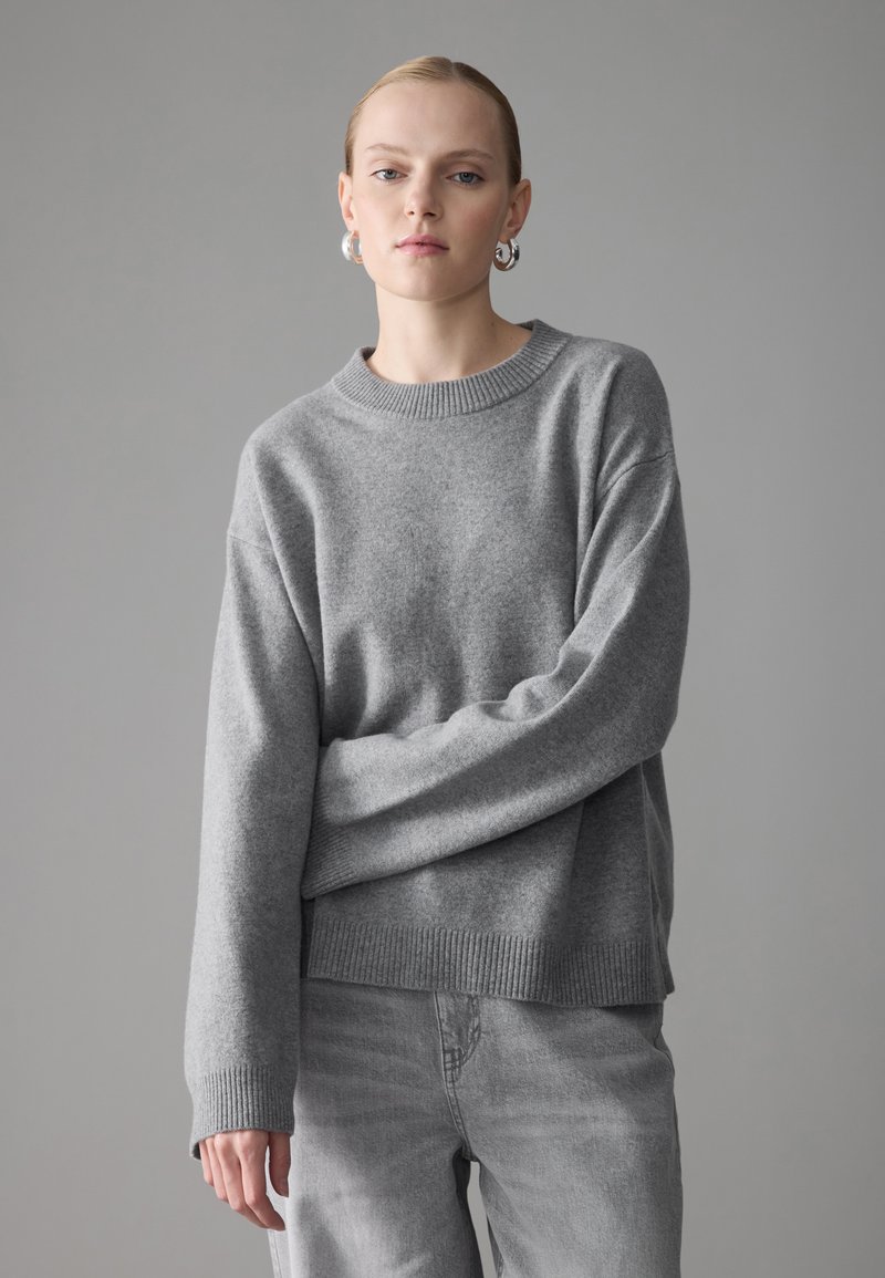 Grauer Pullover mit geripptem Halsausschnitt und Bündchen, aus weicher Wolle gefertigt, hat einen lockeren Schnitt und lange Ärmel, kombiniert mit hellgrauen Jeans.