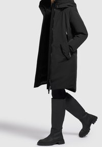 Cappotto lungo nero con cappuccio, dotato di zip frontale e tasche laterali. Abbinato a stivali in gomma al ginocchio con suole texturizzate.