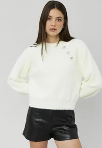 Pull blanc et duveteux à col rond et épaules tombantes, orné de trois accents argentés à paillettes sur le devant. Associé à un short noir texturé.