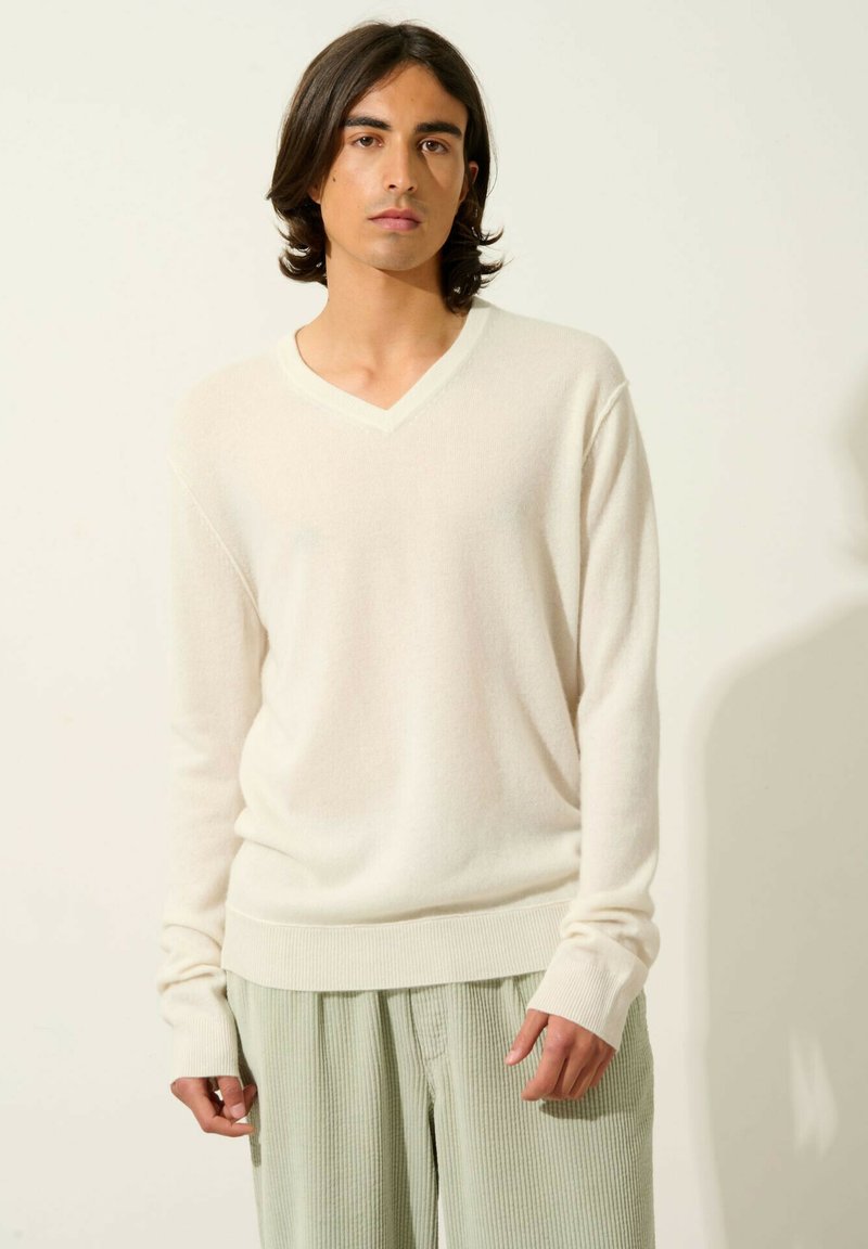 Maglione crema a V in morbido tessuto a maglia, con polsini e orlo a costine, abbinato a pantaloni verdi chiari con texture. Design semplice e moderno.