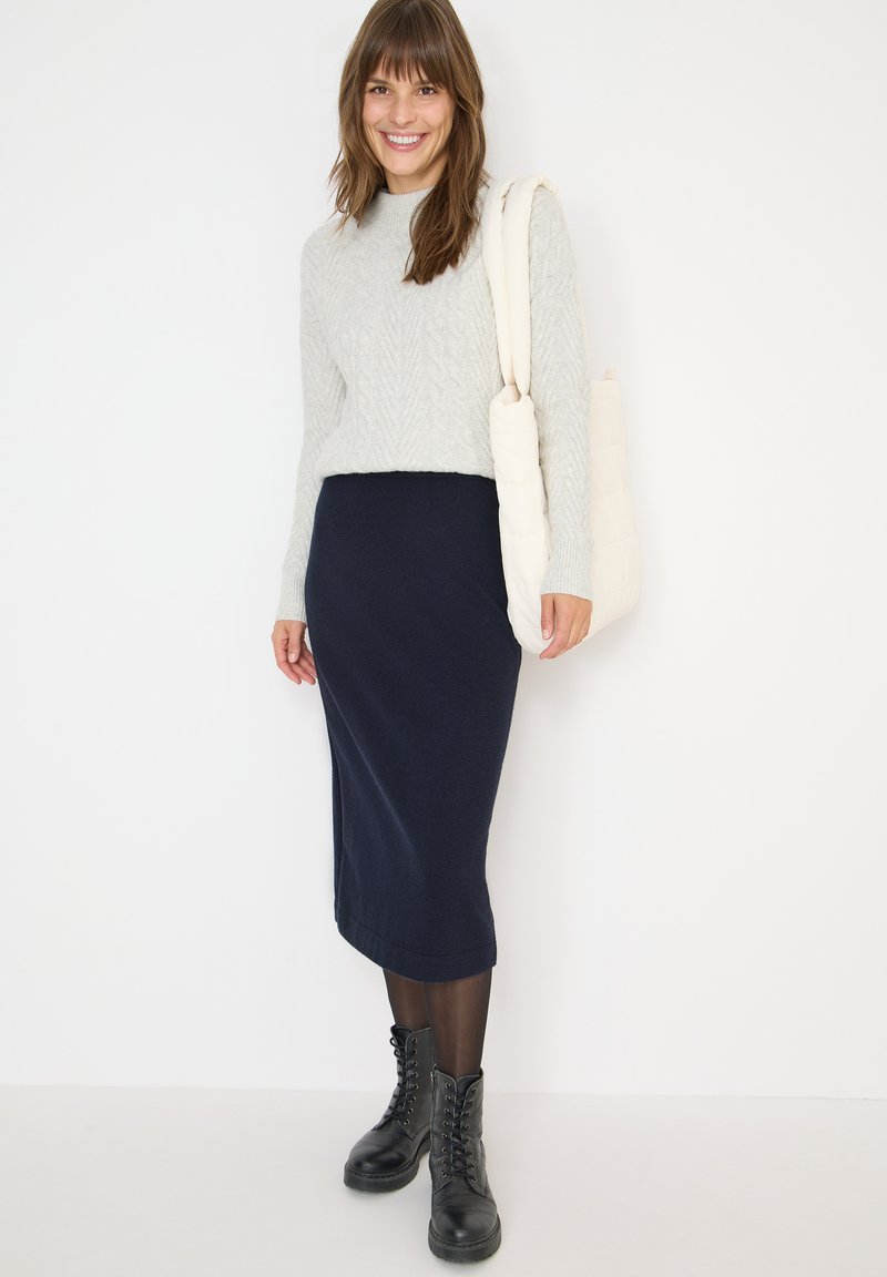 Femme portant un pull gris clair, une jupe midi marine, des bottes noires à lacets, et portant un sac à bandoulière en cuir crème matelassé, souriant devant un mur blanc.