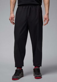 Zwarte sportbroek met een elastische tailleband, voorzien van gestructureerde accenten aan de zijkanten en een taps toelopende pasvorm. G gedragen met zwarte en rode sneakers.