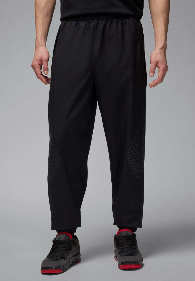 Zwarte sportbroek met een elastische tailleband, voorzien van gestructureerde accenten aan de zijkanten en een taps toelopende pasvorm. G gedragen met zwarte en rode sneakers.