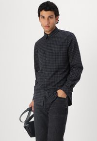 Námornícky károvaná košeľa na gombíky so slim fit strihom, dlhými rukávmi a vreckom. Model má na sebe tmavé džínsy a drží malú čiernu tašku.