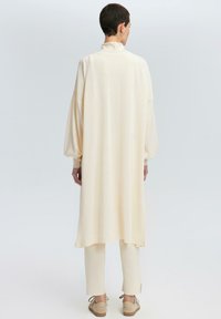 Tunic lunga e oversize crema con colletto alto, maniche a palloncino e polsini a coste, abbinata a pantaloni tapered coordinati e scarpe leggere.