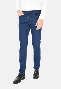 Uomo che indossa jeans slim-fit blu scuro, camicia bianca con bottoni e scarpe eleganti in pelle nera, in piedi con una mano in tasca.