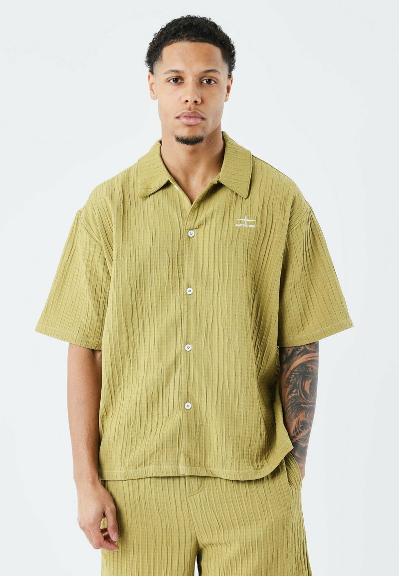 Chemise à manches courtes, verte olive, avec un motif texturé, fermeture à boutons et coupe légèrement surdimensionnée. Présente un logo brodé sur la poitrine gauche.