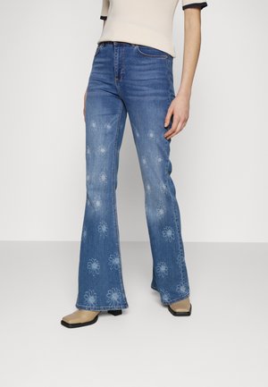 Woman wearing flared blue jeans with faded white floral prints, beige square-toe boots, and a light beige short sleeve top.