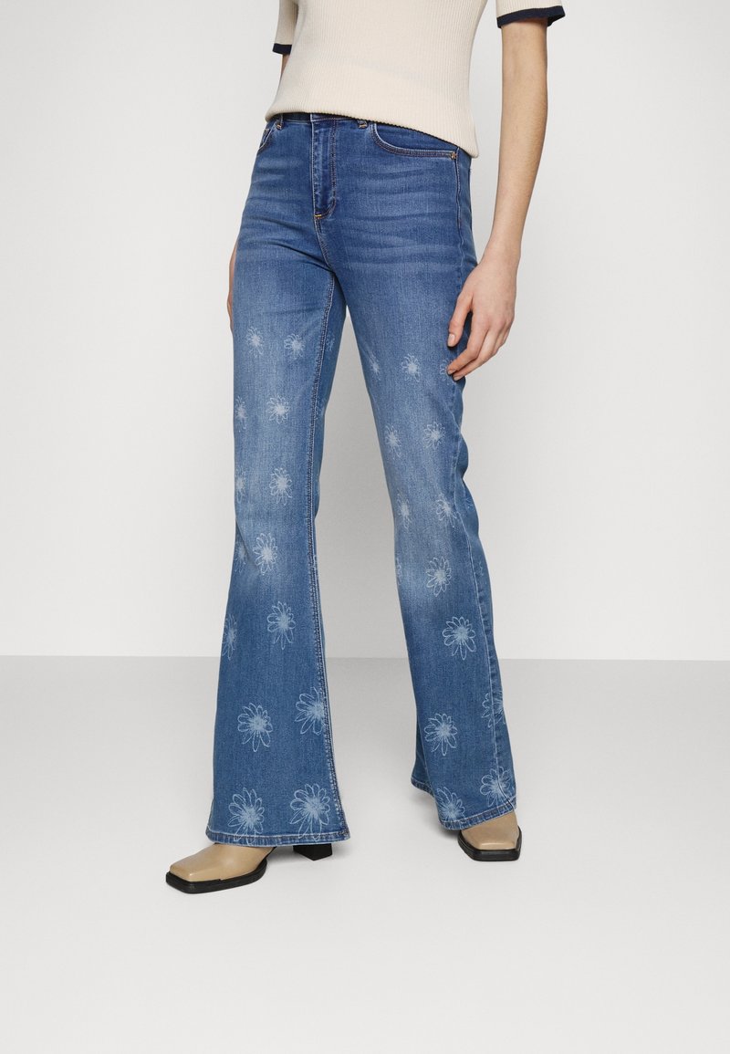 Vrouw in blauwe flare jeans met vervaagde witte bloemenprints, beige vierkante laarzen met korte teen en een lichtbeige T-shirt met korte mouwen.