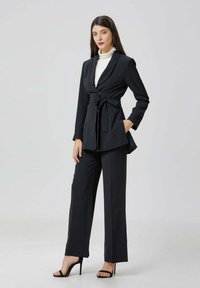 Femme portant un blazer noir ceinturé sur un col roulé blanc, un pantalon noir à jambes larges et des sandales à talons hauts noires, se tenant avec une main dans la poche.