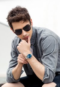 Orologio da polso in rete argentata con quadrante blu e indicatori bianchi. Il modello indossa una camicia grigia con bottoni e occhiali da sole neri, seduto con le dita appoggiate sul mento.