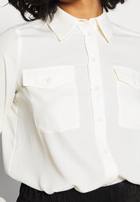 Blouse blanche en tissu lisse, avec un col pointu, une patte de boutonnage à l'avant et deux poches poitrine à rabat.