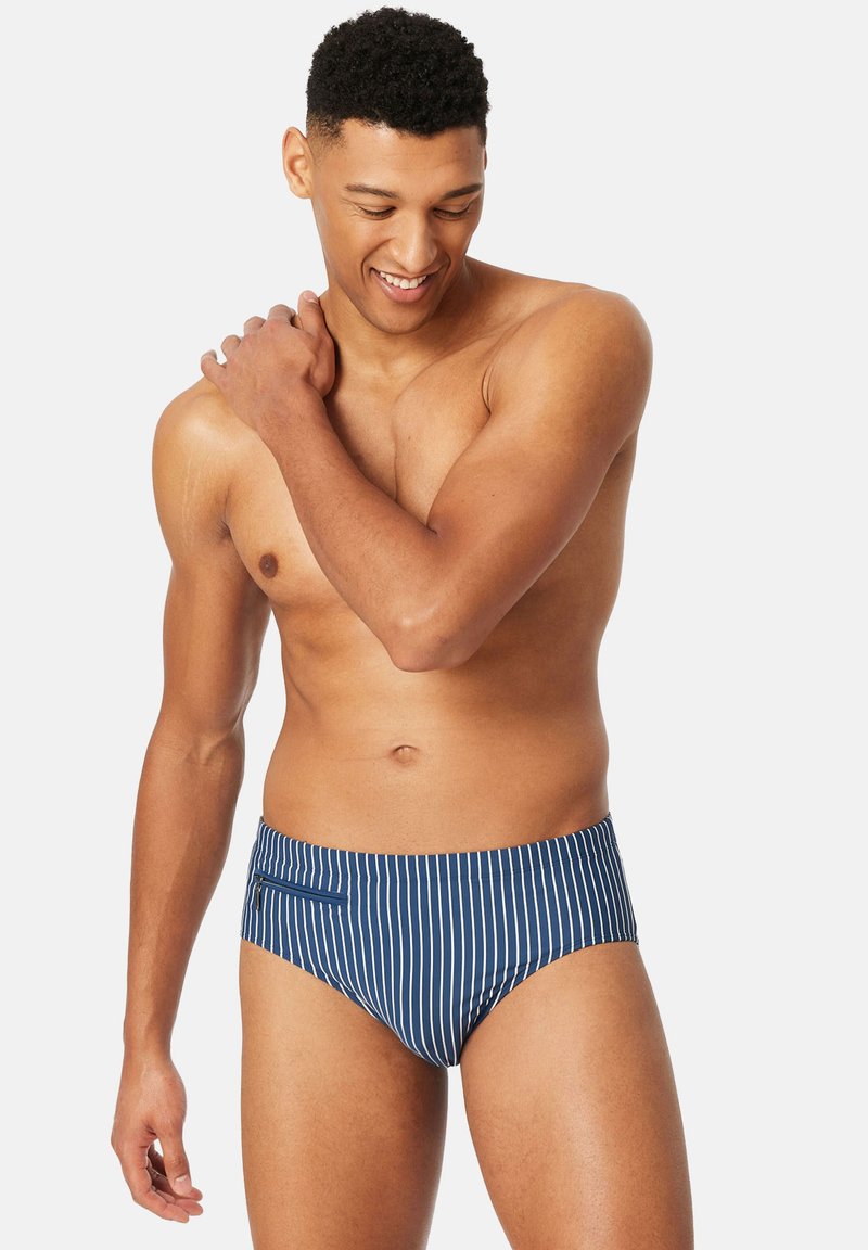 Homme souriant torse nu portant un maillot de bain rayé vertical bleu et blanc avec une petite poche zippée, touchant son épaule gauche.