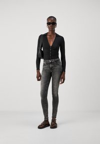 Bodysuit negro de manga larga con escote en V y detalles de botones, combinado con jeans ajustados grises y sandalias negras con tiras. Fondo liso.