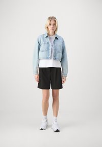Giacca in denim cropped blu chiaro con chiusura a bottoni, due tasche frontali, t-shirt bianca sotto, pantaloni corti neri e scarpe sportive bianche.