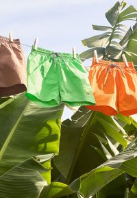 Tre paia di pantaloni corti appesi a una corda: marrone, verde e arancione. Il tessuto è leggero con una vita elastica e dettagli con cordino.