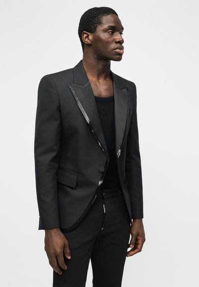 Άνδρας με μαύρο σακάκι με σατινέ πέτα πάνω από μαύρο πουκάμισο και μαύρο παντελόνι, στέκεται μπροστά σε απλό φωτεινό φόντο.
