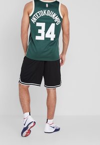 Maglia da basket verde con lettering bianco "ANTETOKOUNMPO" e numero "34", abbinata a pantaloni corti neri con rifiniture bianche e scarpe da ginnastica.