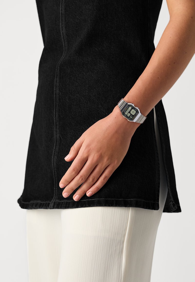 Tunique en denim noir avec une fente latérale, associée à un pantalon blanc texturé. Montre numérique métallique au poignet avec un cadran rectangulaire.