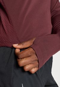 Langärmliges, kastanienbraunes Sportoberteil mit einer strukturierten Einlage, eng anliegenden Bündchen mit Daumenlöchern, kombiniert mit schwarzen Shorts. Nahaufnahme einer Handgriff.