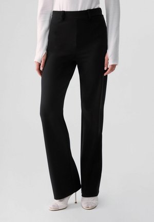 Mujer con pantalones negros de corte recto, camiseta blanca de manga larga y tacones blancos abiertos, de pie frente a un fondo claro y sencillo.