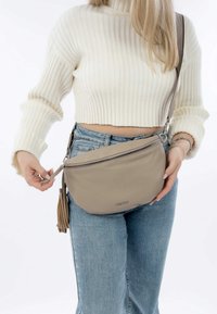 Beige Leder-Crossbody-Tasche mit Reißverschluss, Quaste und glatter Textur, getragen an der Hüfte, während eine Hand des Models den Reißverschluss greift.
