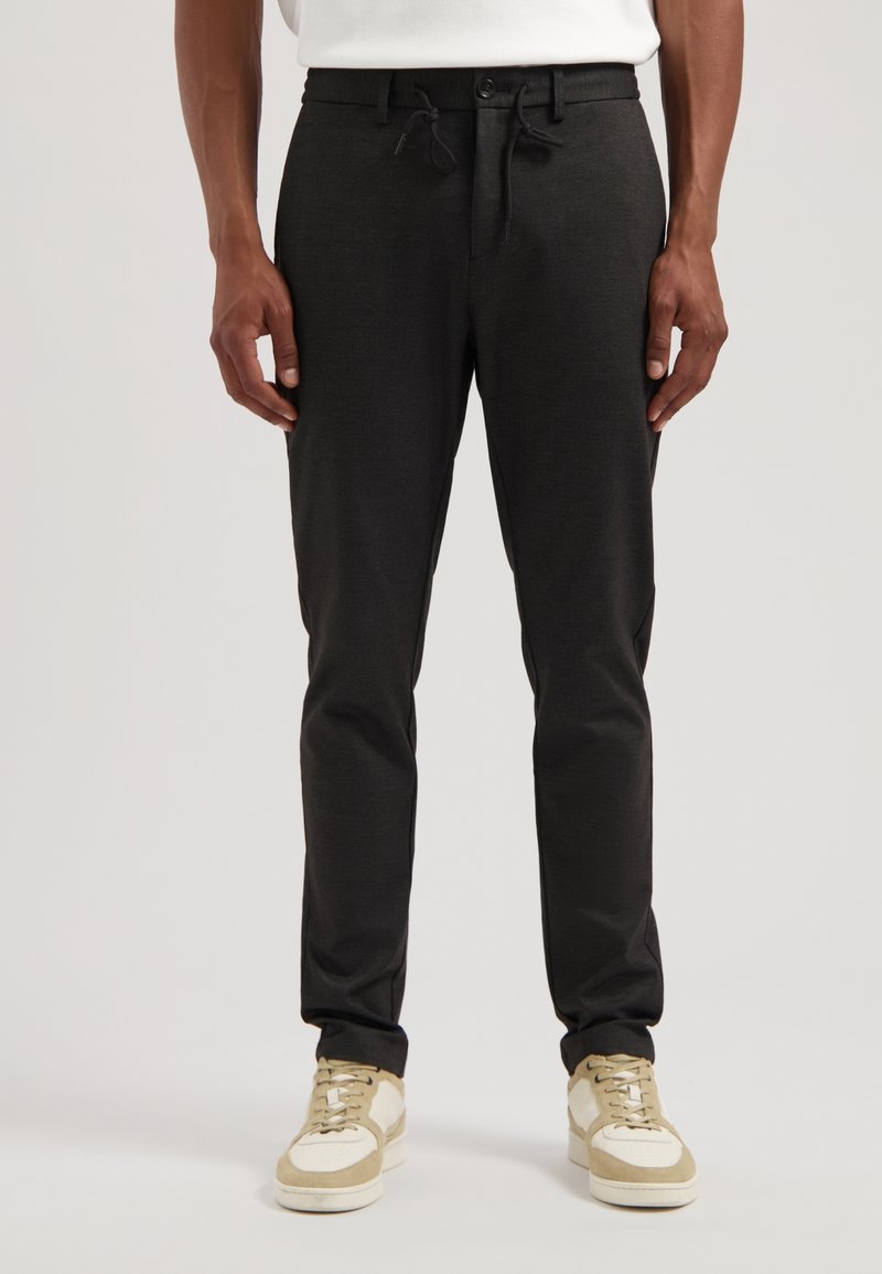 Zwarte, slim-fit broek van een gestructureerde stof, met een trekkoord en knoopsluiting, zijzakken, gecombineerd met beige en witte sneakers.