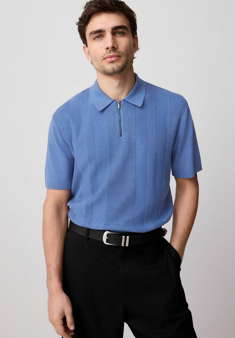 Polo shirt azzurro con maniche corte e zip, caratterizzato da strisce verticali testurizzate, indossato con pantaloni neri e una cintura.
