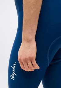 Κοντινό πλάνο του χεριού ενός ατόμου που ακουμπάει δίπλα στο μηρό του, φορόντας σκούρο μπλε ποδηλατικά σορτς με το λευκό λογότυπο "Rapha.".