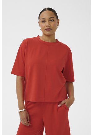 Žena s tamnom kosom vezanom unatrag, nosi široku crvenu majicu kratkih rukava i odgovarajuće crvene hlače, jedna ruka u džepu, a na zapešću srebrna narukvica.