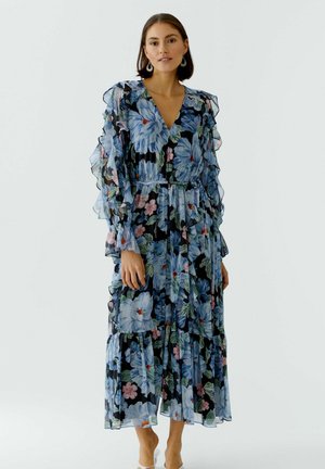 Frau trägt ein langes schwarzes Kleid mit blauen und pinken Blumenmustern und durchsichtigen, gerafften Ärmeln, steht vor einem schlichten Hintergrund.