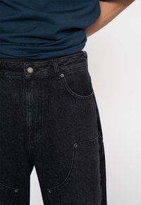 Schwarze Jeans aus Denim mit hoher Taille, vorderen und hinteren Taschen sowie silbernen Hardware-Details. Strukturierter Stoff mit leichtem Glanz.