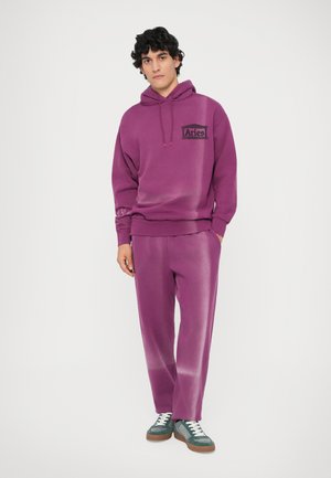 Paarse hoodie met een logo aan de voorkant, bijpassende paarse broek en groene sneakers met witte veters enbruine zolen. Zacht materiaal, ontspannen pasvorm.