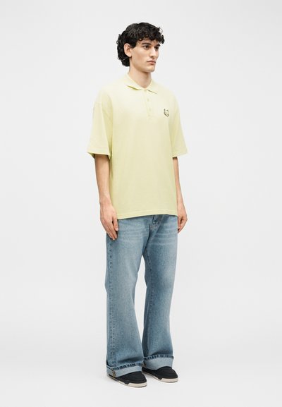 Νεαρός άνδρας στέκεται, φοράει ανοιχτό κίτρινο polo με κοντά μανίκια και φαρδιά μπλε τζιν με μαύρα αθλητικά παπούτσια, σε ένα απλό λευκό φόντο.