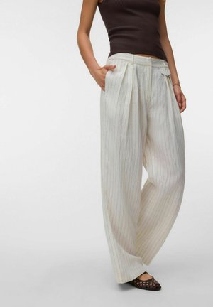 Femme portant un pantalon large blanc à fines rayures, un haut sans manches foncé et des ballerines tissées noires, debout avec une main dans la poche.