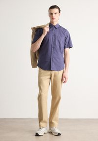 Kortermet, knappet skjorte i dyp lilla med brystlomme, paret med khaki bukser og grå sneakers, holder en beige jakke.