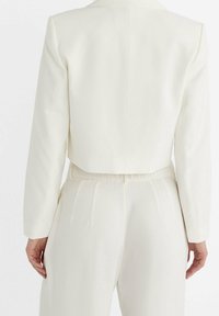 Blazer crème court au design structuré, manches longues et texture lisse ; associé à un pantalon taille haute avec des plis.