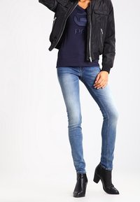 Persona vistiendo jeans skinny de color azul claro, botas de taco negro hasta el tobillo, una camisa azul marino y una chaqueta bomber negra, posando con una mano en la cadera.