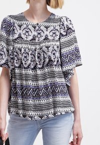 Blusa a maniche corte con intricate fantasie in bianco e nero, dettagli viola, colletto rotondo e particolare arricciato lungo il busto, abbinata a jeans chiari.