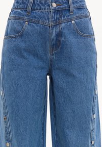 Jean en denim bleu avec poches avant, fermeture par boutons et boutons-pression argentés courant le long des coutures latérales extérieures des jambes.