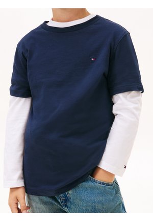 Dziecko ubrane w nałożony na siebie granatowy krótki T-shirt z krótkim rękawem na białą koszulkę z długim rękawem, niebieskie dżinsy, ręka w kieszeni, kadr obejmujący ramiona i biodra.