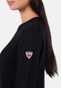 Schwarzer Strickpullover mit rundem Halsausschnitt und gerippten Details, mit einem bunten "Rossignol"-Logopatch auf dem linken Ärmel.