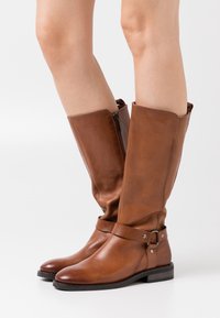 Botas de cuero marrón hasta la rodilla con suela plana, que cuentan con una cremallera lateral y una correa decorativa con un hebilla plateada.