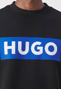 Felpa nera con un'importante toppa rettangolare blu con la parola "HUGO" in grassetto bianco. Tessuto morbido con scollatura rotonda.