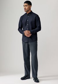 Camicia blu marina con bottoni in tessuto intrecciato, indossata con pantaloni grigi su misura e scarpe nere. Presenta un piccolo logo rosso sul petto.