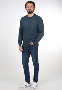 Donkergroene trui met ronde hals en rood logo op de borst, gecombineerd met blauwe spijkerbroek en witte sneakers.