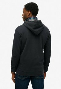 Sudadera negra con capucha, con yugo trasero y puños acanalados, sobre una camisa de franela de cuadros en azul y verde, combinada con jeans oscuros.
