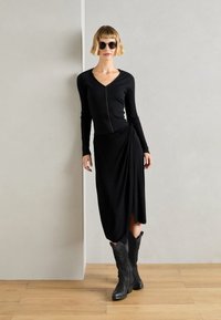 Haut noir à manches longues côtelé avec une fermeture éclair à l'avant, associé à une jupe noire drapée et des bottes noires jusqu'aux genoux. Des lunettes de soleil complètent le look.
