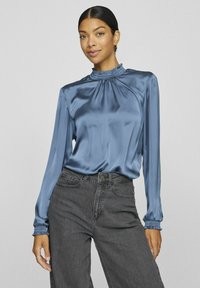 Blusa di satin blu con collo alto arricciato e maniche lunghe. Presenta polsini elasticizzati e una struttura liscia, abbinata a jeans neri a vita alta.