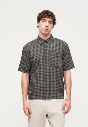 Młody mężczyzna w czarno-białej kratkowanej koszuli z krótkim rękawem i jasnobeżowych spodniach, stojący na jednolitym jasnym tle.